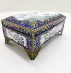 Caixa chinesa em esmalte cloisonné policromado  séc. XX. Elegante caixa retangular em cobre esmaltado cloisonné, ricamente ornamentada com decoração policromada sobre fundo branco e azul-cobalto. A tampa apresenta delicada cena com aves entre flores e ramos de cerejeira, emoldurada por frisos dourados de arabescos e volutas em relevo. As laterais são decoradas com motivos florais estilizados e a peça repousa sobre pés rendilhados em metal dourado. Interior e base esmaltados em branco. Trabalho artesanal de grande refinamento técnico e cromático, típico da tradição chinesa de esmaltação cloisonné. Dimensões: 6 x 16 x 10 centímetros.