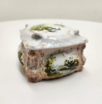 Caixa de formato quadrangular com tampa ajustada, em porcelana moldada e esmaltada, decorada com pinturas manuais de paisagens campestres nas laterais e na tampa, envoltas por arabescos dourados em relevo. Apresenta bordas sinuosas e decoração rocaille, típica do gosto Rococó que marcou a produção de porcelanas alemãs e austríacas no final do século XIX. Interior e base esmaltados em branco. Peça de fino trabalho artesanal, representativa da tradição decorativa da Europa Central. Dimensões: 9 x 13 x 13 centímetros.