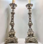 Magnífico par de tocheiros sacros de grande porte, executados em metal banhado a prata, ricamente cinzelados e fundidos em alto-relevo. Apresentam decoração exuberante em estilo barroco, com profusão de volutas, folhas de acanto e elementos fitomórficos, repousando sobre bases triangulares ornadas com volutas. Cada face exibe imagens em relevo de Jesus Cristo, Nossa Senhora e São José, emolduradas por cartelas e motivos rocaille, destacando a inspiração litúrgica e o trabalho minucioso do metal. Corpos torneados e articulados, sustentando bobeches trabalhadas. Peças de grande expressividade artística e devoção, próprias para altares, capelas ou  altares domésticos. Dimensões:1,30 x 35 centímetros.