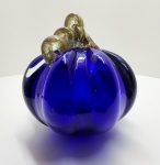 Elegante escultura em vidro artístico soprado, em formato de abóbora estilizada, de corpo facetado e tom azul cobalto profundo, com aplicação superior em vidro retorcido de tonalidade azul escuro translúcido. A peça revela domínio técnico excepcional na manipulação do vidro ainda em fusão, evidenciado na simetria das gomos, no brilho intenso e na pureza do vidro. O contraste entre o corpo liso e a alça retorcida cria um efeito visual vibrante, típico das criações modernas inspiradas na escola Murano, onde o vidro é simultaneamente matéria escultórica e luminosa. O azul cobalto  uma das cores mais difíceis de obter com transparência e uniformidade  é aqui explorado em toda sua profundidade, resultando em uma obra de grande presença visual e sofisticação cromática. Altura: 17 x 17 centímetros. Estado de conservação: Apresenta pequena perda na ramagem.