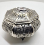 Elegante caixa de formato octogonal executada em metal prateado, repousando sobre pés de apoio ornamentados. A superfície apresenta ricamente gravados motivos fitomórficos em relevo, com destaque para folhas estilizadas em padrão simétrico, típicos do estilo neoclássico tardio / art nouveau de influência europeia. Na tampa, medalhão central oval em porcelana esmaltada com delicada pintura floral em tons de marrom, amarelo e branco, realçando o caráter artesanal da peça. Interior liso, com leve pátina do tempo que realça a autenticidade.Dimensões:10 x 16 centímetros.