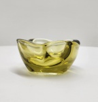 Elegante bowl em vidro artístico de Murano, de forma orgânica com bordas suavemente onduladas, confeccionado em vidro maciço translúcido na tonalidade âmbar dourada. A peça revela leve variação cromática conforme a espessura do vidro, destacando o trabalho artesanal de sopro livre e o refinado polimento manual. Dimensões: 16x8 centímetros.