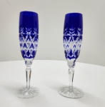 Par de refinadas taças em cristal overlay, com camada externa azul cobalto sobre cristal transparente, lapidadas à mão em padrão geométrico e vegetal estilizado. As hastes facetadas e bases estreladas realçam a luminosidade e o brilho característicos da cristalaria boêmia, símbolo de tradição e excelência artesanal. Dimensões:23x6 centímetros.