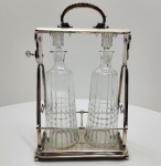 Sofisticado tantalus (porte-liqueur) de fabricação WMF  Württembergische Metallwarenfabrik, Alemanha, c. 1900, composto por duas garrafas em cristal lapidado abrigadas em uma estrutura metálica prateada de elegante design geométrico. A armação, de forma trapezoidal, apresenta alça superior decorada em torção, pés esféricos e o característico mecanismo lateral com chave funcional, que permite travar as garrafas, garantindo segurança e exclusividade  uma das marcas dos modelos de prestígio da WMF.As garrafas, em cristal facetado de alta pureza, exibem lapidação em faixas verticais e tampas facetadas, em perfeita harmonia com o brilho acetinado do metal prateado. Na base, punções WMF gravadas, incluindo o losango com avestruz (símbolo da manufatura) e o código I/O, indicando prata eletrolítica de alta qualidade. Dimensões: ( 38 x 23 x 13 centímetros)