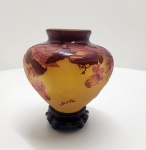 - GALLÉ - Elegante vaso em vidro multicamadas, elaborado pela técnica de vidro camafeu com ácido, característica maior da Escola de Nancy. O corpo em tom âmbar dourado recebe sobreposição em vermelho-rubino e violeta, magistralmente gravada com motivos florais naturalistas, em modelagem profunda e de excelente definição  típico da fase madura de Gallé. A paleta quente, o relevo vigoroso das folhas e a delicadeza dos botões revelam a combinação de virtuosismo técnico e poesia botânica que consagrou Gallé como um dos grandes mestres do Art Nouveau francês. Dimensões: 15x16 centímetros.