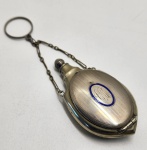 Finíssima flaconette de bolso, Alemanha ou Áustria, período Belle Époque / Art Nouveau, circa 19001915.Frasco feminino de formato achatado e oval, típico das peças de perfumaria portátil utilizadas em chatelaines, bolsas de mão e acessórios de vestuário do início do século XX. Corpo em metal prateado (alpaca / níquel-prata) com superfície decorada em guilloché linear, técnica característica da ourivesaria germânica da época.No centro, medalhão oval em esmalte azul translúcido, com delicado monograma caligrafado sob a camada vítrea. A tampa roscada conserva o acabamento original, com trabalho fino e transição suave entre as linhas gravadas. Acompanha a corrente antiga, igualmente em metal prateado, destinada ao uso em chatelaine.Na base, marcação em relevo GES. GESCHÜTZT, indicação de desenho protegido por lei (modelo registrado), utilizada em território germânico até meados da década de 1940, não correspondendo a fabricante específico. Dimensões:7x4 centimetros.