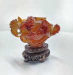 Incensário chinês tripé, inteiramente esculpido em ágata natural, com base original em madeira entalhada. China, c. 19501980.Pequeno vaso trípode de excelente qualidade, talhado em ágata verdadeira de tom âmbar translúcido, com estrias internas e variações naturais típicas da carnelian. O material apresenta brilho vítreo e forte translucidez quando exposto à luz, confirmando a autenticidade da pedra e distinguindo-a do jade e de resinas. A peça exibe alças laterais em forma de dragões ou qilin, habilmente vazadas, e superfície decorada em baixo-relevo. A tampa segue o mesmo padrão de acabamento, e o conjunto repousa sobre base chinesa tradicional em madeira escura, delicadamente entalhada, perfeitamente ajustada ao formato do incensário. Trabalho de notável refinamento dentro da produção chinesa em hardstone da segunda metade do século XX, com boa simetria, polimento fino e excelente presença visual. Dimensões: 11x11x7 centímetros. Apresenta pequenos desgastes e perdas em pontas mais delicadas, comuns em esculturas de pedra. dimensões: 11x11x7 centímetros.