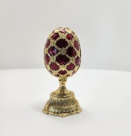 Elegante ovo decorativo no estilo Fabergé, adquirido na Rússia e elaborado em liga metálica de qualidade com banho de ouro. Seu exterior é revestido com esmalte translúcido em tom rubi imperial, aplicado sobre base texturizada em padrão guilloché, criando profundidade e reflexos típicos das peças inspiradas na joalheria imperial russa. A superfície exibe arabescos e elementos folheados em ouro, formando uma treliça rica em detalhes. A abertura articulada revela interior esmaltado em rubi profundo. A peça apresenta duas surpresas, seguindo a tradição Fabergé. Dimensões: 12x6 centímetros.