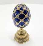 Sofisticado ovo decorativo no estilo Fabergé, adquirido na Rússia e confeccionado em liga metálica banhada a ouro, com esmaltação translúcida em tom azul safira profundo. O acabamento guilloché sob o esmalte cria efeitos de luz característicos das peças inspiradas nos ovos imperiais produzidos para a corte dos Romanov. A superfície externa apresenta treliças douradas em relevo, com ornamentos florais e pequenos cristais decorativos, formando uma composição clássica do estilo Russo Imperial. Abertura articulada revela interior esmaltado em azul claro, com padrões florais em relevo. No centro, a peça traz duas surpresas tradicionais: Mini relógio funcional em estilo relógio de bolso, com tampa esmaltada. Mini coroa imperial cravejada, também banhada a ouro, decorada com cristais cintilantes.