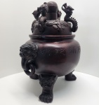 Magnífico incensário chinês de grande porte, executado em cerâmica de alta temperatura com acabamento em tonalidade castanho-escura, imitando o aspecto do bronze tradicional. O corpo globular apresenta inscrições devocionais em chinês, associadas à proteção e purificação espiritual. As laterais são adornadas por imponentes cabeças de leões guardiões (Shi/Fu Lion) que funcionam como alças decorativas. A peça se apoia sobre três pés em forma de patas de leão, reforçando a simbologia protetiva ligada à cultura chinesa. A tampa, ricamente modelada, apresenta dragões ascendentes em alto-relevo, elementos ligados ao poder, prosperidade e energia vital. Dimensões: 60x53x40 centímetros.