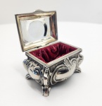Elegante caixa decorativa em metal prateado, trabalhada em estilo Rococó com folhas, volutas e motivos florais em alto-relevo. O tampo apresenta composição central emoldurada com flores e ornamentos em estilo europeu clássico. As laterais exibem relevos ondulados e pés recurvados, típicos da estética Rocaille. O interior é revestido em veludo vermelho. Dimensões:5x 7 x 4 centímetros.