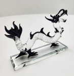 Escultura representando um dragão estilizado, modelado à mão em vidro artístico soprado. O corpo serpenteado em vidro cristal transparente exibe textura escamada, enquanto detalhes como garras, crina, bigodes e cauda são aplicados em vidro preto, criando forte contraste visual. A peça repousa sobre base retangular em vidro lapidado, conferindo estabilidade e elegância à composição. O dragão, símbolo de força, proteção e prosperidade nas culturas asiáticas, torna esta escultura ideal para decoração de ambientes. Dimensões:10x14x4centímetros.