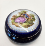 Elegante caixa redonda em porcelana fina de Limoges, França, decorada com esmaltação em bleu de four (azul cobalto profundo) e rica douração em ouro polido. A tampa apresenta uma tradicional cena galante ao estilo de Fragonard, finamente pintada à mão, com grande vivacidade de cor e delicadeza nos detalhes. A borda é ornamentada com guirlandas e arabescos dourados, aplicados manualmente, técnica característica dos ateliês decoradores de Limoges do século XX. Interior em porcelana branca, brilho impecável. Dimensões:6x1centímetros.