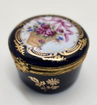 Encantadora caixa de porcelana Limoges Castel France, comercializada pela H. Stern, em esmalte azul-cobalto profundo com decoração de ouro 22k aplicada à mão. A tampa exibe um cesto de flores em policromia delicada, envolvido por arabescos dourados finamente delineados, que ressaltam o brilho do esmalte vítreo. As ferragens de bronze patinado mantêm o estilo tradicional das caixas francesas de luxo, conferindo charme e solidez ao conjunto. O fundo apresenta a marca Limoges Castel France e indicação de ouro 22k, atestando a qualidade superior da manufatura. Dimensões 3x5 centímetros.