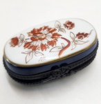 Caixinha francesa Limoges, peça inteiramente pintada à mão. Modelo oval em porcelana fina, com corpo em azul profundo e tampa esmaltada em branco, decorada com flores em tom sépia-avermelhado e contornos dourados delicados. A peça apresenta acabamento em metal trabalhado e fecho frontal ornamentado, mantendo o estilo tradicional das caixas de Limoges. Dimensões: 3x4x6 centímetros.
