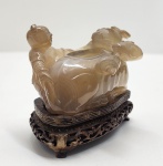 Elegante escultura chinesa talhada em ágata natural translúcida, representando uma criança apoiada sobre um boi reclinado  um tema auspicioso associado à prosperidade, serenidade e à harmonia entre o homem e a natureza. A pedra apresenta tonalidade mel com belas variações internas, típicas da ágata de boa qualidade utilizada em peças decorativas finas do período tardo-Qing a início da República (c. 18801930). Dimensões: 9x10x6 centímetros. Apresenta pequenos desgastes e perdas em pontas mais delicadas, comuns em esculturas de pedra.