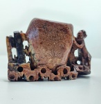 Escultura oriental em pedra jaspeada de tonalidades castanho-avermelhadas, habilmente talhada à mão, representando um macaco em postura ativa apoiado sobre rochas orgânicas enquanto sustenta um recipiente de estúdio. A superfície exibe veios naturais do jaspe, com variações cromáticas que criam profundidade e reforçam o caráter mineral da peça. O recipiente central apresenta entalhes florais delicados, enquanto a base, recortada em formas irregulares, integra cavidades naturais do próprio jaspe, valorizando a estética bruta e artesanal. A figura do macaco  símbolo de inteligência, astúcia e proteção na iconografia oriental. Dimensões: 7x11x3 centímetros.  Apresenta pequenos desgastes e perdas em pontas mais delicadas, comuns em esculturas de pedra.