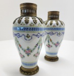 Elegante par de vasos franceses em porcelana fina da manufatura real de Sèvres, datados de 1771 conforme a marca de letra-data S pintada em azul sob o esmalte. As peças apresentam corpo ovóide delicadamente pintado à mão com guirlandas florais, laços azulados e festões de rosas em policromia suave, característicos da estética refinada de meados do século XVIII. Os vasos são montados em bronze dourado (ormolu) com aplicações de folhas estilizadas junto ao gargalo e bases circulares com frisos decorativos, reforçando o luxo típico das encomendas aristocráticas do período pré-Revolução. A paleta clara, a pincelada minuciosa e o equilíbrio das proporções evidenciam o trabalho de ateliês especializados da manufatura em sua fase áurea. Conjunto raro, harmonioso e absolutamente representativo do estilo Louis XVI. Dimensões: 20x10 centímetros cada.