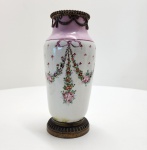 Elegante vaso em porcelana Sèvres com pintura esmaltada em tons de rosa, lilás e verde, apresentando guirlandas florais pendentes, laços estilizados e ramos pontilhados típicos do repertório decorativo de Sèvres. A peça possui montagens originais em bronze dourado (ormolu) no topo e na base, trabalhadas com motivos de perolado e festões. O bojo exibe delicado trabalho pictórico manual e esmaltação de alta qualidade. No fundo, marca pintada em azul sob o vidrado, correspondendo ao monograma de Sèvres. Dimensões: 23x12 centímetros.  ( Apresenta restaurações).