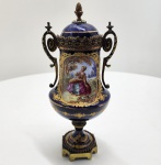 Elegante urna decorativa em porcelana de Paris, França, século XIX, com tampa original e montagens em bronze dourado (ormolu).Peça em porcelana esmaltada no clássico bleu roi (azul-real), ricamente ornamentada com douração aplicada a pincel, apresentando volutas, guirlandas e arabescos em ouro. Na face principal, medalhão oval pintado à mão com sena galante em ambiente campestre, executada com grande delicadeza cromática. Alças laterais de desenho sinuoso em bronze, bem como base quadrada facetada, também em bronze dourado. A tampa segue o mesmo padrão decorativo, com pináculo em forma de botão. Dimensões: 26x11 centímetros ( tampa esta colada)