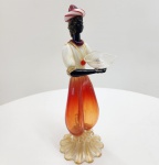 Elegante escultura Blackamoor em vidro de Murano, inteiramente modelada à mão pelos mestres vidreiros italianos. A peça apresenta figura masculina estilizada, apoiada sobre base em forma de pétalas e tonalidades âmbar e rubi. O corpo alongado em vidro soprado, os braços em vidro preto opalino e o turbante bicolor demonstram a complexidade das técnicas tradicionais de Murano. A escultura mantém o adesivo antigo Made in Italy, típico das produções das ilhas de Murano entre as décadas de 1950 e 1970. Dimensões:41x14 centímetros.