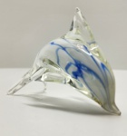 Escultura em vidro artístico representando golfinho em salto, executada em massa de vidro cristalino com inserções ondulantes em tons de azul e branco, criando efeito de movimento fluido e transparências elegantes. A peça apresenta modelagem orgânica, nadadeiras delineadas e corpo suavemente torcido, demonstrando domínio da técnica de vidro soprado. Dimensões: 8x12x7 centímetros.