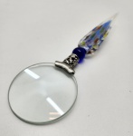 Lupa artística com cabo em vidro Murano, peça contemporânea em execução manual, apresentando cabo alongado em vidro soprado com aplicações de murrine multicoloridas e aventurina, terminando em esferóide azul profundo unido a ferragem decorada em metal prateado trabalhado; lente circular de amplo diâmetro, transparente e de boa graduação, formando conjunto elegante que combina utilidade e arte em um objeto refinado de caráter decorativo e colecionável. Dimensões: 20x6 centímetros.