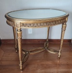 Elegante mesa de aparador em madeira entalhada, trabalhada em autêntico estilo Luís XVI, ricamente dourada e decorada com profusão de elementos clássicos. O tampo semicircular repousa sobre uma cinta ornamentada por frisos contínuos de acantos, guirlandas estilizadas, ondas volutadas e molduras peroladas  motivos característicos do repertório decorativo neoclássico francês do final do século XVIII.A peça apoia-se sobre quatro pés torneados e canelados em espiral (típicos do estilo), com nós ornamentados por florões. A base é unida por travessa em forma de elipse quadrifólia, com acabamento perolado e flor central, garantindo estabilidade e reforçando a sofisticação do desenho. Sob a cinta, há guirlandas florais pendentes, finamente esculpidas e douradas, acentuando o aspecto cerimonial da mesa. O conjunto apresenta douração com pátina, preservando brilho e profundidade no relevo.78x90x65 centímetros.