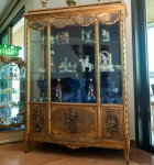 Imponente vitrine em madeira entalhada, executada em estilo Luís XVI, com frontaria envidraçada e rica decoração em douração. A parte superior apresenta friso contínuo de folhas de louro, sobre o qual se destacam arcos moldurados sustentados por guirlandas florais pendentes, laços e rosetas  elementos característicos do repertório neoclássico francês do final do século XVIII.As portas em vidro possuem molduras ornadas por entalhes helicoidais e ferragens em metal lavrado, mantendo o desenho clássico da peça. O interior é equipado com prateleiras de vidro espesso, permitindo excelente visibilidade para objetos de coleção e cristalaria. A parte inferior é fechada por painéis em palhinha trançada, sobre os quais surgem apliques entalhados de tochas, coroas e guirlandas, reforçando o caráter cerimonial e decorativo típico das peças de inspiração cortesã. Os pés cabriolet discretos completam a estrutura, com ornamentos florais e acabamento dourado com pátina suave. Dimensões: 1,80x1,34x0,80.