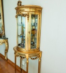 Delicada vitrine estilo Luís XVI, executada em madeira entalhada e ricamente dourada. O corpo frontal apresenta vidros curvos e molduras trabalhadas com frisos perolados, folhas de louro e caneluras, elementos clássicos do repertório neoclássico francês. O topo é ornamentado por um coroamento de rosas e laços, sobre friso de folhas entrelaçadas. A faixa superior é decorada com festões florais suspensos, finalizados por rosetas e palmetas. A parte inferior da vitrine forma um pequeno aparador semicircular, sustentado por quatro pés longos e canelados, unidos por guirlandas florais em relevo. Ao centro destaca-se um medalhão circular com perfil neoclássico, ladeado por ornamentos vazados e guirlandas pendentes. O interior conta com prateleiras de vidro e fundo espelhado, conferindo luminosidade e realce às peças expostas. A douração apresenta pátina quente e desgaste artístico, preservando o caráter decorativo. Dimensões:1,80x0,75x0,40 metros.