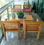 Conjunto composto por quatro cadeiras artesanais e uma mesa de centro maciça, produzidos em madeira tropical pesada (como teca, acácia ou similar), com acabamento natural e entalhes manuais característicos do estilo indo-colonial/balinês. As cadeiras apresentam braços recurvos ricamente entalhados, encosto em ripas horizontais com travessas cruzadas e medalhão central, além de assentos amplos com almofadas em tecido vermelho. As pernas são torneadas e reforçadas, garantindo estabilidade e durabilidade. A mesa de centro, inteiramente em madeira maciça, possui pés largos em curva descendente ("pés em escora") e tampo espesso, próprio de mobiliário artesanal asiático. O desenho harmoniza com as cadeiras formando um conjunto único, elegante e robusto. Dimensões: mesa 0,41x1,24x0,74, cadeiras 0,82x0,62x0,50.