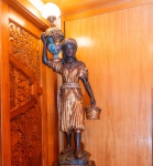Escultura Blackamoor veneziana, esculpida em madeira maciça e ricamente policromada, representando figura feminina em trajes exóticos, carregando jarro ornamentado com frutos, do qual se eleva uma luminária com cúpula em vidro opalino. A peça apresenta entalhe manual característico: cabelos, vestes, frutas e ornamentos em alto relevo, com aplicação de pátina dourada e policromia brilhante, finalizada com verniz protetor. O corpo é pintado em tom ébano, conforme tradição dos blackamoors venezianos dos séculos XVIIIXIX.A figura apoia-se sobre base rocosa esculpida, instalada sobre pedestal octogonal em madeira, também patinado e decorado em tons terrosos. Trata-se de peça de grande presença, ideal para halls, escadarias e ambientes clássicos.