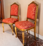 Par de cadeiras em madeira entalhada e dourada, com trabalho minucioso em folhas de acanto, laços e frisos geométricos, características associadas ao estilo Louis XVI. Molduras com pátina dourada original, apresentando desgaste natural que reforça o caráter decorativo e a autenticidade do conjunto. Encostos em formato levemente arqueado, com coroamento decorado por laço entalhado. Assentos amplos e confortáveis, revestidos em tecido brocado vermelho de padrão floral clássico. Pernas dianteiras curvas (cabriolet) e traseiras levemente arqueadas, ambas com entalhes decorativos.
