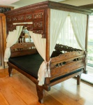 Imponente cama balinesa antiga em madeira maciça, ricamente entalhada à mão, apresentando estrutura tradicional de dossel e composição arquitetônica típica das ilhas da Indonésia. A peça exibe um extraordinário repertório decorativo, com frisos, painéis vazados e esculturas zoomórficas e florais distribuídas por toda a extensão da cabeceira, laterais e coroamento superior.Os entalhes revelam trabalho artesanal de grande profundidade e refinamento, destacando motivos de folhagens estilizadas, flores tropicais e criaturas mitológicas, elementos característicos do repertório artístico balinês. Os pés apresentam forma escultural espiralada, com talha volumosa e acabamento original, preservando pátina antiga e marcas do tempo coerentes com sua idade e origem.O dossel mantém a moldura integral com frisos entalhados e acompanha cortinados em tecido leve, que reforçam a estética exótica e cerimonial da peça. Dimensões: 2,10x2,45x1,34. (Desmontar e retirada do apartamento por conta  do arrematante)