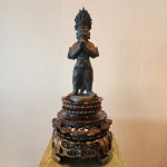 Escultura balinesa em bronze, representando figura protetora (penjaga), com postura de saudação ritual. Peça de tradição hindu-balinesa, com coroa flamejante e traços rústicos característicos da arte votiva da ilha. Acompanha base em madeira entalhada (não original). Bali, início a meados do século XX.