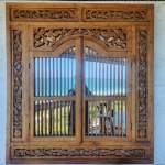 Espelho janela balinesa em madeira maciça, ricamente entalhada à mão. A moldura apresenta painéis laterais, barra superior e friso inferior trabalhados com motivos florais e folhagens sinuosas, em clássica linguagem ornamental da marcenaria tradicional de Bali.A parte central com folhas articuladas de gradeado vertical.Dimensões: 1,30x1,20 metros.