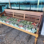 Banco longo em madeira maciça, com encosto trabalhado em painéis vazados geométricos e entrelaçados, formando desenhos lineares que equilibram leveza e robustez. Os braços são levemente curvos, suavizados por volutas discretas. As pernas frontais são torneadas, com frisos verticais e anéis.Assento acompanhado por almofadão estofado em tecido floral tropical, acrescentando conforto e função decorativa. Dimensões:0,87x1,84x0,54.