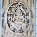 Placa decorativa em madeira maciça, trabalhada em entalhe vazado, representando Ganesha, divindade hindu associada à sabedoria e à prosperidade. A figura aparece sentada em posição tradicional, com múltiplos braços segurando atributos simbólicos, cercada por folhas e flores de lótus em composição oval.O entalhe apresenta bom volume, detalhes nas vestes, joias e ornamentos da figura, com acabamento patinado em tom claro que ressalta o relevo. A peça encontra-se montada sobre painel de fibra trançada que funciona como suporte decorativo.Dimensões: escultura 38x30 centímetros.