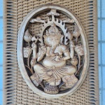 Placa decorativa em madeira maciça, trabalhada em entalhe vazado, representando Ganesha, divindade hindu associada à sabedoria e à prosperidade. A figura aparece sentada em posição tradicional, com múltiplos braços segurando atributos simbólicos, cercada por folhas e flores de lótus em composição oval.O entalhe apresenta bom volume, detalhes nas vestes, joias e ornamentos da figura, com acabamento patinado em tom claro que ressalta o relevo. A peça encontra-se montada sobre painel de fibra trançada que funciona como suporte decorativo.Dimensões: escultura 38x30 centímetros.