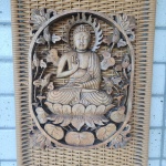 Placa decorativa em madeira maciça, entalhada em alto relevo, representando Buda sentado em flor de lótus, rodeado por ramos floridos em composição vazada de formato oval. A figura apresenta auréola flamejante, vestes detalhadas e expressão serena, seguindo iconografia tradicional do budismo do Sudeste Asiático.O conjunto encontra-se montado sobre painel retangular de fibra trançada.Dimensões sem painel: 38x29 centímetros.