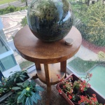 Mesa lateral redonda em madeira maciça, produzida artesanalmente em Bali. O tampo, de corte espesso, exibe veios naturais bem marcados, característicos de madeiras tropicais da região. A peça possui três pernas curvas, unidas por um prateleira circular inferior, reforçando a estrutura e oferecendo funcionalidade adicional. Dimensões: 62x49 centímetros.