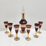 Conjunto composto por licoreira e seis taças em cristal turco, trabalhado em técnica overlay rubi (camada de cristal colorido sobre base transparente) com rica aplicação de ouro e esmaltação decorativa. A licoreira apresenta stopper facetado em cristal transparente e corpo em rubi intenso, ornamentado com faixas largas de ouro polido e arabescos em esmalte branco com detalhes pontilhados.As seis taças seguem o mesmo padrão decorativo, com bojo rubi e hastes completamente revestidas em ouro, além de bases decoradas com motivos em esmalte e ouro, típicos da produção turca