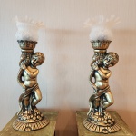 Conjunto formado por duas luminárias ornamentais executadas em bronze patinado, cada peça modelada com a figura de um putto em pose graciosa, abraçado a uma coluna de inspiração clássica, com rica textura e detalhes minuciosamente esculpidos.As luminárias são coroadas por globos em vidro leitoso opalino, com delicado relevo de uvas e folhas, finalizados por bordas onduladas que suavizam a luminosidade quando acesas. A composição remete à estética neoclássica / romântica, com forte apelo decorativo e presença cênica. ornamentais executadas em bronze patinado, cada peça modelada com a figura de um putto em pose graciosa, abraçado a uma coluna de inspiração clássica, com rica textura e detalhes minuciosamente esculpidos.. Dimensões: 64x22x15 centímetros.