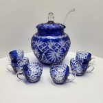 Elegante conjunto de ponche produzido em cristal overlay azul cobalto, proveniente da tradicional escola vidreira da Boêmia (Alemanha). A poncheira apresenta corpo globular de paredes espessas, com aplicação de cristal incolor internamente e camada externa azul profundamente lapidada. O trabalho de lapidação revela motivos radiantes, palmetas estilizadas e trama geométrica em losangos cruzados, típicos da produção boêmia do final do século XX.Acompanha tampa original, com pegador facetado de recorte prismático, além de concha em cristal soprado. O conjunto inclui seis xícaras correspondentes, também em overlay azul, com rica lapidação frontal e alças em cristal totalmente incolor, mostrando o contraste de tonalidades e transparências característico da técnica. Dimensões 35x26 centímetros.