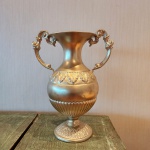 Vaso decorativo em metal com acabamento dourado, apresentando formato clássico com corpo ovóide, bojo ornamentado por friso em relevo de folhas estilizadas e base canelada. Alças laterais simétricas, em volutas decorativas, reforçam a estética tradicional. Peça de presença, com brilho homogêneo e excelente apelo decorativo. Dimensões: 32x26x16 cencentímetros.
