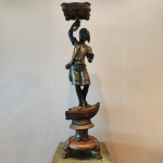 Importante Blackmoor veneziano em madeira talhada, policromada e dourada, representando figura masculina erguendo pequeno tabuleiro ornamental acima da cabeça. A escultura apresenta fisionomia marcada, acabamento lacado escuro e vestes ricamente decoradas com motivos florais e arabescos típicos da tradição veneziana. A base, igualmente trabalhada, compõe pedestal de grande presença, apoiado sobre quatro pés zoomórficos. Trabalho elaborado, cenográfico e característico da produção decorativa veneziana entre o final do século XIX e início do XX, quando Blackmoors eram criados como elementos de luxo para salões e palácios. Pátina antiga bem preservada e excelente presença escultórica. Dimensões: 0,95 x 022 x 0,22 centímetros.