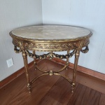 Mesa auxiliar oval em madeira dourada ao gosto Luís XVI, tampo em mármoreElegante mesa de formato oval, trabalhada em madeira ricamente entalhada e dourada, apresentando friso com motivos entrelaçados e reservas florais. A cintura é ornamentada por guirlandas aplicadas, unidas aos montantes torneados e estriados, terminando em pés afunilados com anéis decorativos. Estrutura inferior com travessão arqueado e elemento central em forma de pinha. Tampo em mármore claro, de belíssimo veio natural, perfeitamente ajustado à borda moldurada. Peça de refinado caráter decorativo, inspirada nos modelos franceses do final do século XVIII.