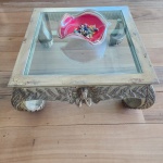 Mesa de centro de proporções robustas, executada em madeira ricamente entalhada com folhas estilizadas ao longo de toda a saia e dos pés. No centro de cada face, destaca-se mascarão de querubim moldado em alto-relevo, conferindo caráter decorativo singular. Apresenta acabamento patinado em tom cru envelhecido, com craquelê acentuado e desgastes voluntários que reforçam o aspecto antigo da peça. Tampo em vidro plano embutido, permitindo a visualização do interior e valorizando o trabalho escultórico periférico. Dimensões 0,44x1,05x1,05.