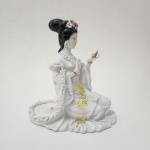 Figura feminina oriental em porcelana esmaltada, trabalho chinês de meados do século XX, ricamente decorada com flores aplicadas e detalhes pintados em policromia suave. A personagem, ajoelhada, segura uma pequena flor, com vestes cuidadosamente modeladas e orladas em pasta trabalhada. Dimensões: 29x26x13