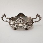 Linda terrina decorativa em metal banhado a prata, exuberantemente trabalhada no estilo rococó, com profusa ornamentação em relevo composta por volutas, cartelas e elementos assimétricos de forte dinamismo. Corpo oval apoiado sobre quatro pés modelados, e ladeado por duas asas vazadas de desenho sinuoso, conferindo elegância e movimento ao conjunto. Interior liso e borda recortada, demonstrando refinamento técnico e decorativo. Peça de presença marcante, representativa da ourivesaria decorativa europeia do final do século XIX ao início do XX.